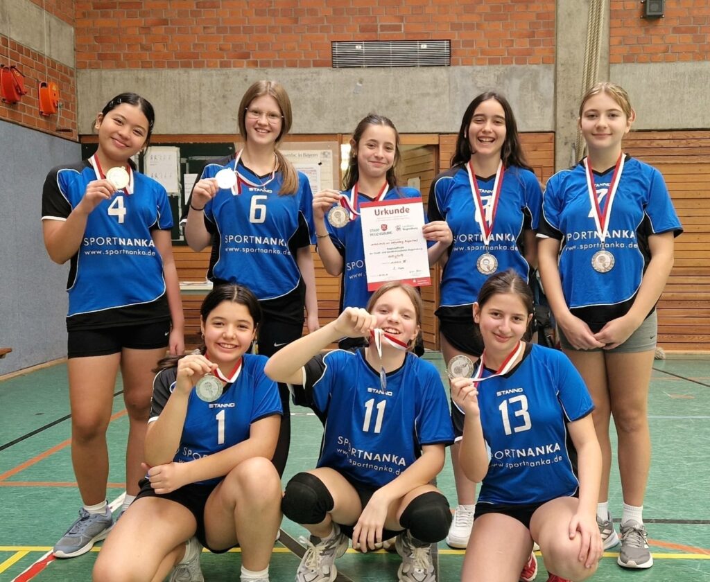 Gruppenfoto der Volleyball-Schulmannschaft in einer Turnhalle: Acht Schülerinnen in blauen Trikots stehen und knien zusammen, tragen Medaillen um den Hals und präsentieren stolz ihre Urkunde für den 2. Platz bei der Regionalmeisterschaft in Regensburg.