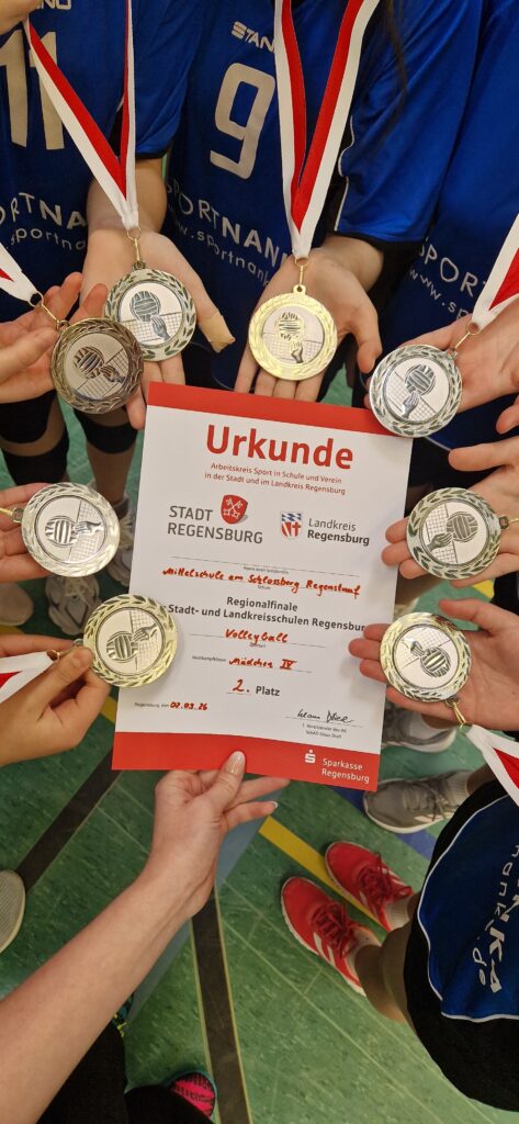 Mehrere Schülerinnen in blauen Volleyballtrikots stehen im Kreis und halten Medaillen sowie eine Urkunde der Regionalmeisterschaft Regensburg in den Händen. Auf der Urkunde ist der 2. Platz im Volleyball vermerkt.